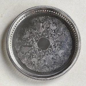 May include: A round silver-toned metal serving tray featuring an intricate engraved floral and scrollwork pattern across the center. The tray has a raised, twisted rope-style border and a perforated rim with small rectangular cutouts.