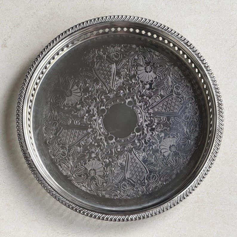 May include: A round silver-toned metal serving tray featuring an intricate engraved floral and scrollwork pattern across the center. The tray has a raised, twisted rope-style border and a perforated rim with small rectangular cutouts.