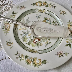 K&ouml;nnte beinhalten: Ein wei&szlig;er Kuchen-Teller mit floralen Mustern in Gelb, Blau und Rot und einem gr&uuml;nen Rand. Ein silberner Tortenheber liegt auf dem Teller. Der Teller und der Heber liegen auf einer wei&szlig;en Tischdecke.