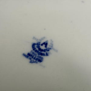 May include: A white ceramic surface featuring a blue stamped maker's mark. The mark includes a central letter A surrounded by a wreath, with the text "WILLOW" and "ENGLAND" printed below, indicating the origin and pattern of the ceramic piece.