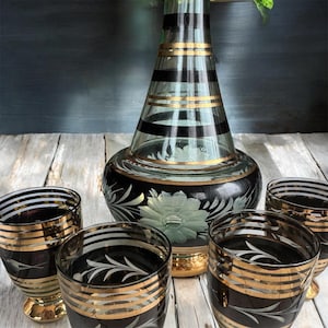 May include: A set of four black and gold striped glass shot glasses with a matching black and gold striped glass decanter with a floral design. The decanter has a clear glass body with black and gold stripes and a floral design.