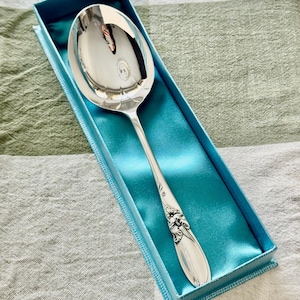 Op de afbeelding: Een zilveren serveerlepel met een decoratief handvat, gepresenteerd in een blauwe geschenkdoos met een satijnen voering. De lepel heeft een grote, ovale kom en sierlijke bloemendetails. De doos is rechthoekig.