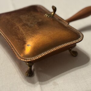 May include: A vintage copper-toned crumb tray with a wooden handle and brass accents. The rectangular tray has decorative trim and rests on small brass feet. The handle is attached with a brass fitting. The tray is designed to collect crumbs.