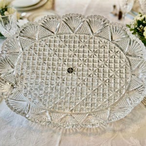 May include: Clear, oval-shaped crystal serving platter with a detailed geometric pattern. The platter features a scalloped edge with fan-like designs. A small, circular emblem is visible in the center. Ideal for serving appetizers or desserts.