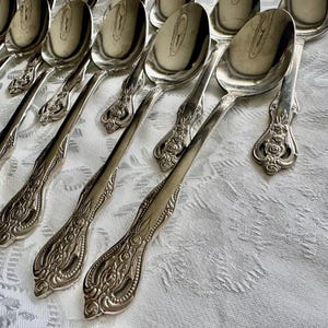 May include: A collection of ornate silver spoons arranged on a white patterned tablecloth. The spoons have detailed floral and scrollwork designs on the handles and a reflective, polished surface.
