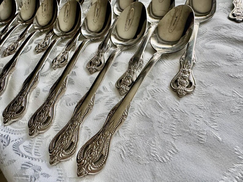 May include: A collection of ornate silver spoons arranged on a white patterned tablecloth. The spoons have detailed floral and scrollwork designs on the handles and a reflective, polished surface.