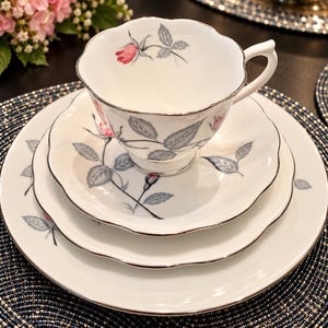 May include: A white porcelain tea set consisting of a teacup, saucer, and plate with scalloped edges and silver trim. The set features a delicate pattern of pink rosebuds and gray leaves on each piece, creating a cohesive floral design.
