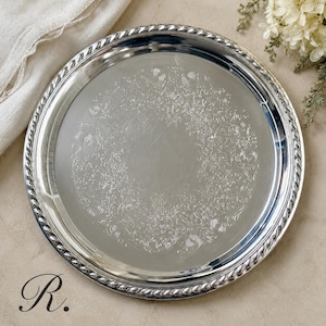 May include: A round silver-toned serving tray featuring a polished center and an intricate floral engraving. The rim is designed with a raised, twisted rope-style border. The surface reflects light, highlighting the detailed etched patterns.