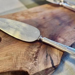 May include: Silver cake server with a decorative handle, resting on a wooden cutting board. The server has a wide, flat blade and a detailed handle. The wood grain of the cutting board is visible.