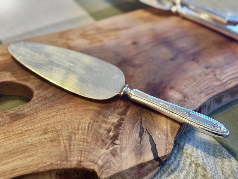 May include: Silver cake server with a decorative handle, resting on a wooden cutting board. The server has a wide, flat blade and a detailed handle. The wood grain of the cutting board is visible.