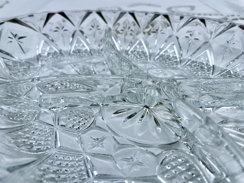 May include: Close-up of a clear, divided glass serving dish. The dish features a detailed, geometric pattern with star and diamond shapes. The glass has a textured surface, reflecting light.