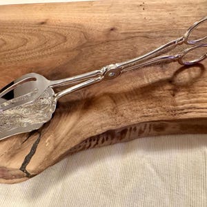 May include: Silver serving tongs with an ornate design on the handles. The tongs are on a wooden surface.