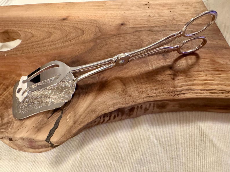 May include: Silver serving tongs with an ornate design on the handles. The tongs are on a wooden surface.