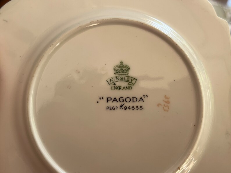 K&ouml;nnte beinhalten: Ein wei&szlig;er Keramikteller mit einem gr&uuml;n-goldenen Logo, auf dem "Ainsley England" und "Pagoda" mit der Nummer "K94635" steht.