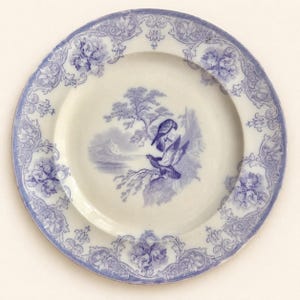 May include: A round ceramic plate with a cream-coloured base and a blue transferware pattern. The centre features a landscape illustration of two birds perched on a branch near a lake. The rim is decorated with a detailed floral and scroll border.