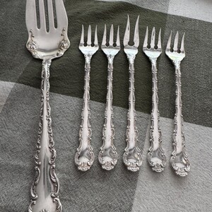 K&ouml;nnte beinhalten: Ein Set aus f&uuml;nf silberfarbenen Metallgabeln mit verzierten, gepr&auml;gten Schn&ouml;rkeln an den Griffen. Die Sammlung umfasst eine gro&szlig;e Vorlegegabel mit vier Zinken und vier kleinere Gabeln, drei davon mit vier Zinken und eine mit zwei Zinken.