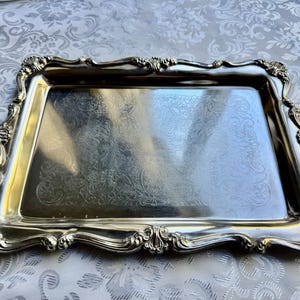 Puede incluir: Una bandeja de servicio rectangular de tono plateado con un borde elevado y ornamentado con volutas intrincadas y motivos de conchas. La superficie central de la bandeja es lisa y reflectante, decorada con un delicado patr&oacute;n floral grabado.
