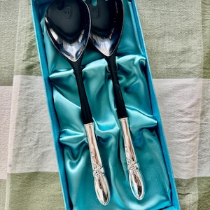Op de afbeelding: Een saladeservingset in een doos met zwarte serveerkoppen en handvatten. De handvatten hebben een zilveren bloemenontwerp aan de basis. De set wordt gepresenteerd in een blauwe doos met een satijnen voering.