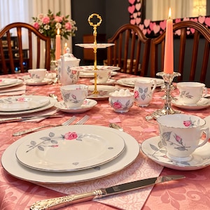 May include: A formal table setting featuring white porcelain dinnerware with a pink rose and grey leaf pattern, silver-toned flatware, a tiered serving stand, a teapot, a sugar bowl, and a tall pink taper candle in a silver holder.