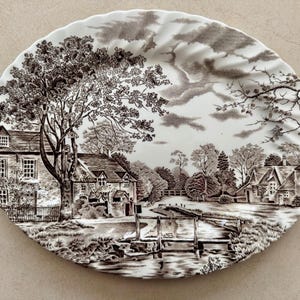 May include: An oval ceramic platter featuring a detailed brown and white transferware print of a rural landscape. The scene depicts stone cottages, a large tree, a small wooden bridge over a stream, and a cloudy sky, all framed by a scalloped edge.