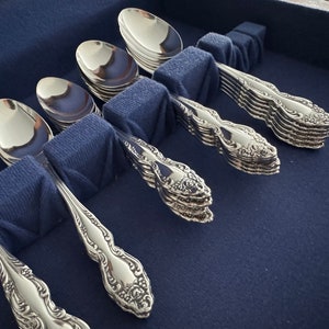May include: A set of polished silverware, including spoons and forks, arranged in a blue velvet-lined case. The silverware has ornate, floral-inspired detailing on the handles. The spoons and forks are neatly organized, reflecting light.