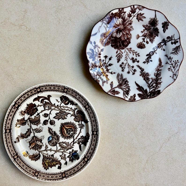 May include: Two ceramic plates with brown floral patterns. One plate features a scalloped edge with a dense botanical print, while the other has a smooth circular rim and a detailed central design with brown, blue, and gold accents.