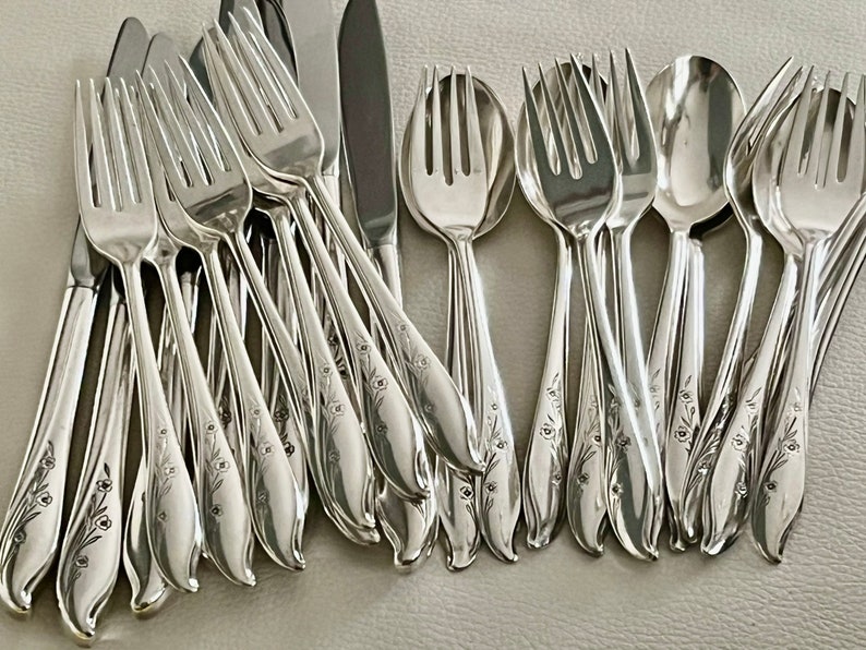 May include: A collection of shiny silver flatware, including forks, spoons, and knives. The silverware has a floral design on the handles. The utensils are arranged in a slightly overlapping pattern, reflecting light.