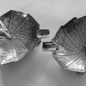 K&ouml;nnte beinhalten: Zwei silberfarbene Aschenbecher, jeweils in Form einer Hand, die ein gef&auml;chertes Kartenspiel h&auml;lt. Die Karten weisen detaillierte Illustrationen auf. Die Aschenbecher haben eine rechteckige Ablage f&uuml;r Zigaretten.