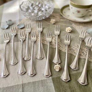May include: An assortment of silver forks with ornate handles, displayed on a green and white checked tablecloth. The forks are alongside a teacup and saucer, small white roses, and a glass bowl filled with sweets.