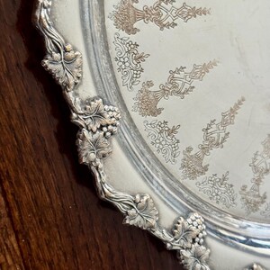 May include: A round silver-toned metal serving tray featuring an ornate raised border decorated with embossed grape clusters and vine leaves. The center of the tray displays a repeating engraved pattern of floral urns and scrollwork.