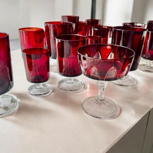 May include: A set of 11 red glass drinking glasses, including a coupe glass, on a white surface. The glasses are all different shapes and sizes, but they are all made of clear glass with a red tint.