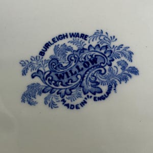 May include: A white ceramic surface featuring a blue transfer-printed maker's mark. The mark displays the text BURLEIGH WARE, WILLOW, and MADE IN ENGLAND, surrounded by an ornate, leafy, and scroll-like decorative border in a deep blue color.
