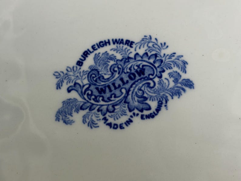 May include: A white ceramic surface featuring a blue transfer-printed maker's mark. The mark displays the text BURLEIGH WARE, WILLOW, and MADE IN ENGLAND, surrounded by an ornate, leafy, and scroll-like decorative border in a deep blue color.
