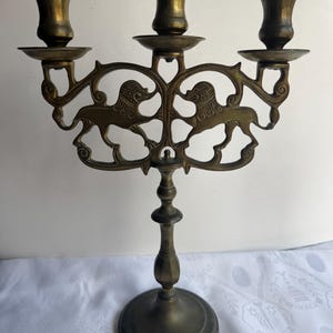 Pu&ograve; includere: Candelabro antico in ottone con tre portacandele. Il candelabro presenta un design ornato con due figure di leoni. La base &egrave; rotonda e lo stelo &egrave; dettagliato. L'altezza totale &egrave; di circa 25 cm.