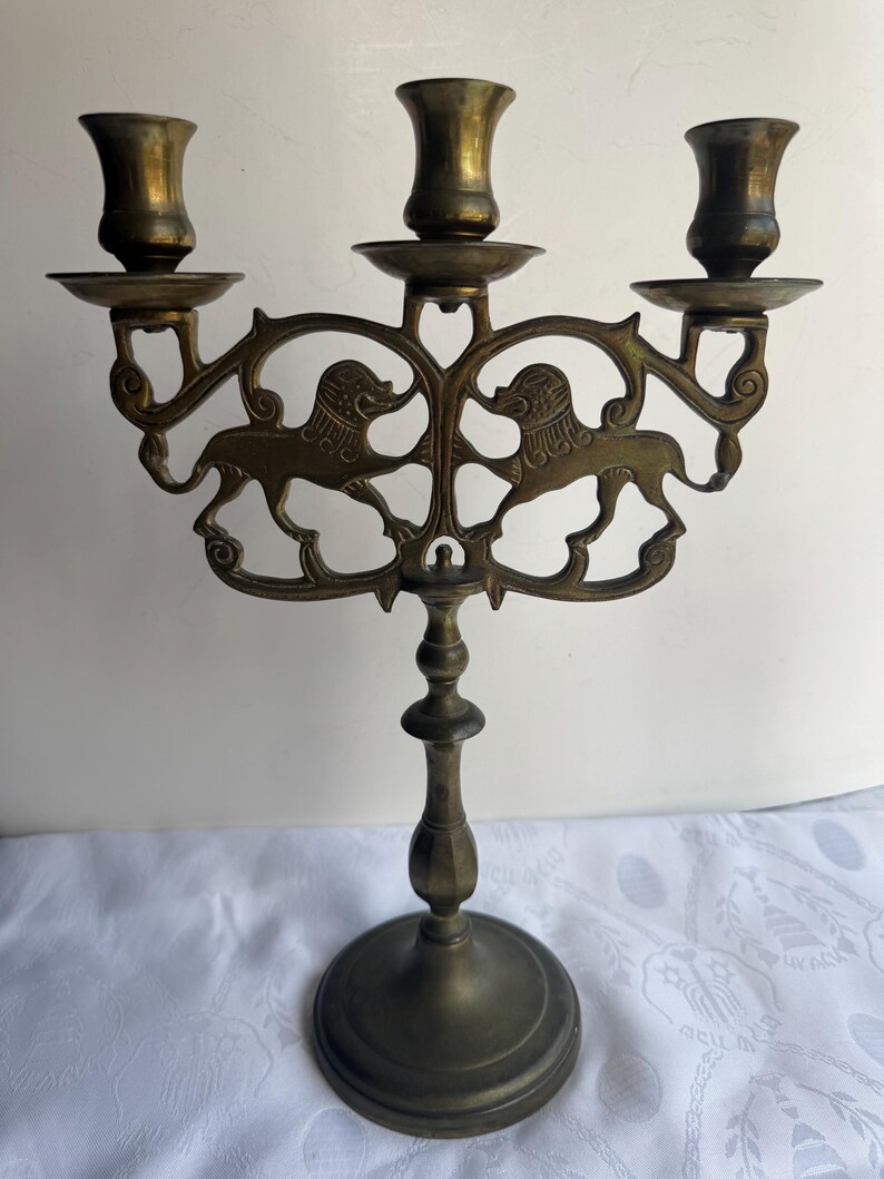 Pu&ograve; includere: Candelabro antico in ottone con tre portacandele. Il candelabro presenta un design ornato con due figure di leoni. La base &egrave; rotonda e lo stelo &egrave; dettagliato. L'altezza totale &egrave; di circa 25 cm.