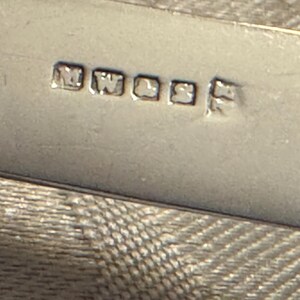 K&ouml;nnte beinhalten: Nahaufnahme eines silbernen Objekts mit den eingestempelten Buchstaben "MWL&S". Das Objekt hat eine reflektierende Oberfl&auml;che und befindet sich vor einem strukturierten Hintergrund. Die Buchstaben sind in einer quadratischen Schriftart.