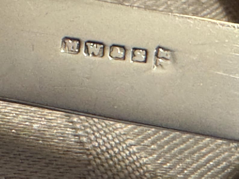 K&ouml;nnte beinhalten: Nahaufnahme eines silbernen Objekts mit den eingestempelten Buchstaben "MWL&S". Das Objekt hat eine reflektierende Oberfl&auml;che und befindet sich vor einem strukturierten Hintergrund. Die Buchstaben sind in einer quadratischen Schriftart.