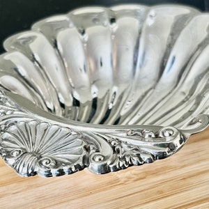 May include: A silver, shell-shaped serving dish with a decorative, ornate rim. The dish is made of metal and has a smooth, reflective surface.
