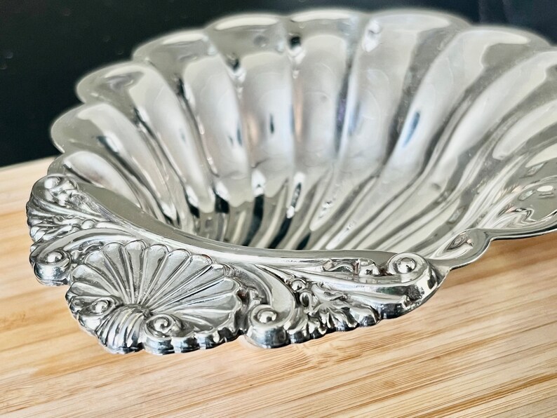 May include: A silver, shell-shaped serving dish with a decorative, ornate rim. The dish is made of metal and has a smooth, reflective surface.