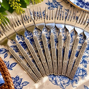 May include: A set of eight stainless steel forks arranged in a fan shape. Each fork features a polished metal head and a rectangular handle with an embossed, intricate scrollwork pattern and small cross-shaped details.