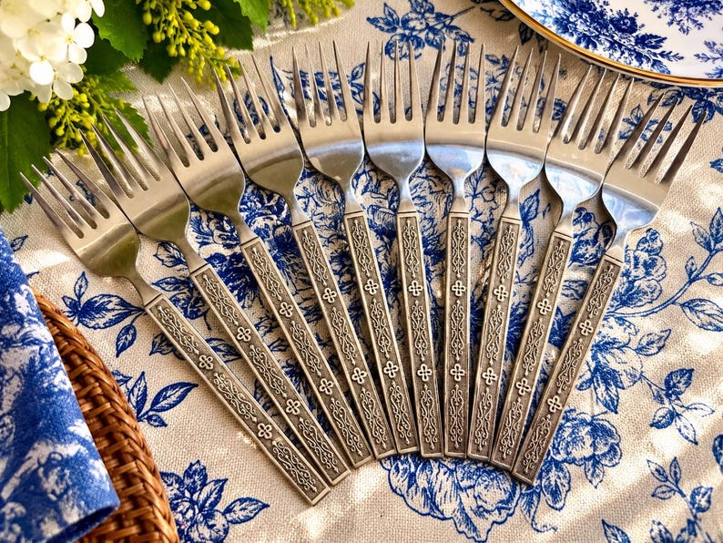 May include: A set of eight stainless steel forks arranged in a fan shape. Each fork features a polished metal head and a rectangular handle with an embossed, intricate scrollwork pattern and small cross-shaped details.