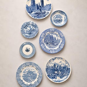 May include: A collection of seven ceramic plates featuring blue and white transferware patterns. The designs include detailed illustrations of historic buildings, landscapes, and architectural scenes, each surrounded by intricate floral borders.