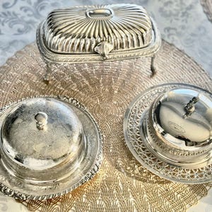 May include: An assortment of antique silver serving dishes. The image features a rectangular lidded butter dish with a fluted design, and two round butter dishes with ornate lids and bases. The dishes are arranged on a decorative placemat.