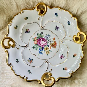 Puede incluir: Plato de porcelana blanco para servir con un centro floral y ribetes dorados. El plato presenta un borde festoneado y asas doradas. El dise&ntilde;o central muestra un ramo de flores rosas, moradas, azules y amarillas.