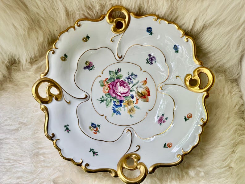 Puede incluir: Plato de porcelana blanco para servir con un centro floral y ribetes dorados. El plato presenta un borde festoneado y asas doradas. El dise&ntilde;o central muestra un ramo de flores rosas, moradas, azules y amarillas.
