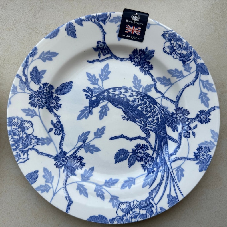 May include: A white ceramic plate featuring a blue transferware pattern of a long-tailed bird perched on flowering branches. A black tag at the top displays the text Royal Wessex, Made in England, and Est. 1795 with a small Union Jack flag icon.
