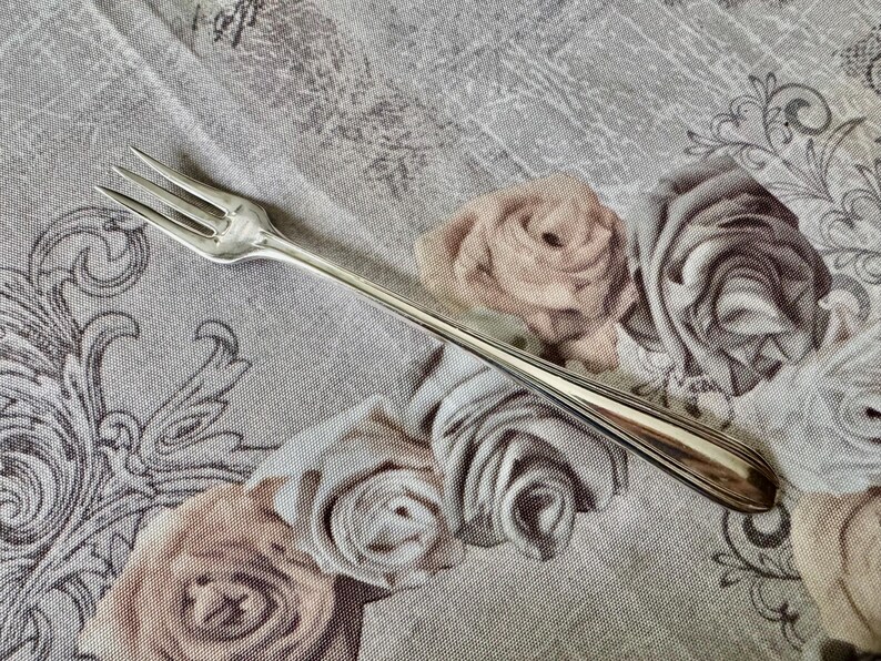 May include: A silver fork with three tines, lying on a floral patterned fabric. The fabric features roses in shades of pink and gray, with decorative scrollwork. The fork has a long, slender handle.