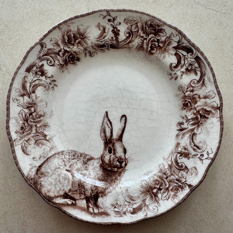 May include: A round ceramic plate with a cream-colored base and a brown crackle glaze finish. The plate features a detailed brown illustration of a rabbit in the lower center and a decorative border of brown floral patterns and scrollwork.