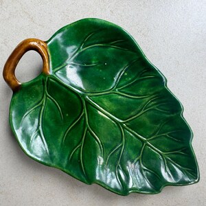 Puede incluir: Un plato de cer&aacute;mica verde brillante con forma de hoja grande y un asa marr&oacute;n. La superficie presenta venas en relieve que imitan la estructura natural de una hoja, con un esmalte liso y reflectante que cubre toda la parte superior.