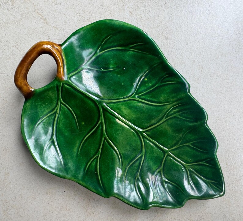 Puede incluir: Un plato de cer&aacute;mica verde brillante con forma de hoja grande y un asa marr&oacute;n. La superficie presenta venas en relieve que imitan la estructura natural de una hoja, con un esmalte liso y reflectante que cubre toda la parte superior.